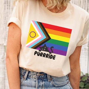 Puede incluir: Una camiseta beige con una bandera arcoíris de orgullo y un gato negro asomándose por detrás. El texto "Purrride" está impreso debajo del gato.