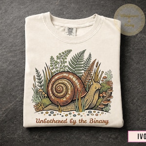 Op de afbeelding: Een ivoorkleurig t-shirt met een grafische illustratie van een bruine slak omringd door groene varens, mos en paddenstoelen. Onder de illustratie staat de tekst "Unbothered by the Binary" in een bruin, afgerond lettertype.