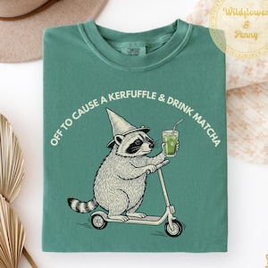 May include: A sage green t-shirt featuring a graphic of a raccoon wearing a pointed hat, riding a scooter, and holding a glass of matcha. The text above the raccoon reads, "OFF TO CAUSE A KERFUFFLE & DRINK MATCHA." The shirt has a crew neckline.