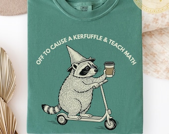 May include: A sage green t-shirt featuring a graphic of a raccoon wearing a pointed hat, riding a scooter, and holding a coffee cup. The text above the raccoon reads, "OFF TO CAUSE A KERFUFFLE & TEACH MATH" in a curved, white, sans-serif font.