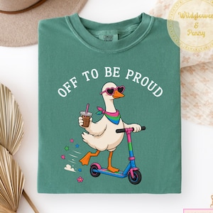 May include: Light green sweatshirt with the text "OFF TO BE PROUD". The graphic features a cartoon goose wearing heart-shaped sunglasses and a scarf, riding a scooter and holding a coffee. The scooter is blue, pink, and green.