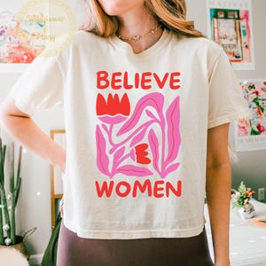 Puede incluir: Una camiseta de manga corta color marfil con el texto rojo "BELIEVE WOMEN" impreso encima y debajo de una ilustración gráfica de dos tulipanes rojos con tallos y hojas rosas. La camiseta tiene un cuello redondo y un corte holgado.