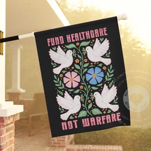 Op de afbeelding: Zwarte vlag met de tekst "FUND HEALTHCARE NOT WARFARE" in roze. De vlag toont witte duiven, roze en blauwe bloemen en groene bladeren. De vlag hangt buiten.