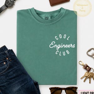 May include: A sage green t-shirt featuring the white text "COOL Engineers CLUB" in a circular design on the chest. The shirt is displayed alongside dark blue denim jeans, a black wristwatch, a brown leather wallet, keys, and sunglasses.
