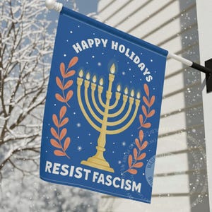 May include: A blue flag with the words "Happy Holidays" and "Resist Fascism" in white. It features a gold menorah and orange leaf accents. The flag is mounted on a white pole against a snowy background.