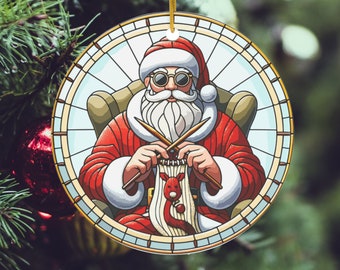 Pode incluir: Enfeite de Natal redondo com o Pai Natal num design de vitral. O Pai Natal, de fato vermelho e óculos, faz tricô de um cachecol vermelho e branco com um desenho de rena. O enfeite é pendurado por um fio dourado.