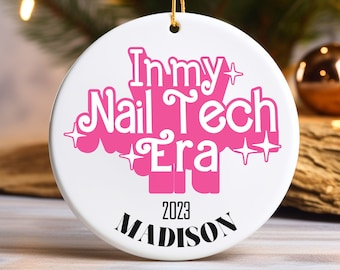May include: A white ceramic Christmas ornament with the text "In my Nail Tech Era" in pink, the year "2023", and the name "Madison" in black. The ornament is round and has a gold string for hanging.