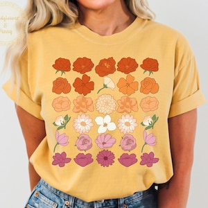 Könnte beinhalten: Senfgelbes T-Shirt mit einem Blumendesign in Orange-, Pfirsich-, Rosa- und Weißtönen. Das Shirt hat einen Rundhalsausschnitt und kurze Ärmel. Der Text "Wildflower & Penny" befindet sich in einem Kreis oben links. Das Wort "MUSTARD" steht in einem rosa Rechteck.