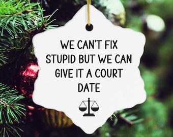 May include: White snowflake-shaped Christmas ornament with the text "WE CAN'T FIX STUPID BUT WE CAN GIVE IT A COURT DATE" in black. A gold string is attached for hanging. The ornament features a scale of justice symbol.