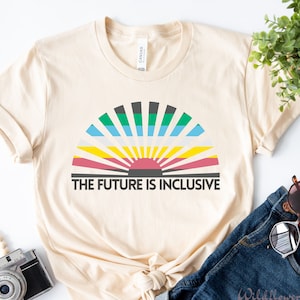 May include: A cream-colored short-sleeve t-shirt featuring a graphic of a semi-circular sunburst with horizontal stripes in black, green, blue, white, yellow, and pink. Below the graphic, the text reads THE FUTURE IS INCLUSIVE in black.