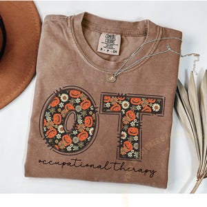 Könnte beinhalten: Braunes Comfort Colors T-Shirt mit den Buchstaben "OT" in einem Kürbis- und Blumendesign. Darunter steht "occupational therapy". Eine silberne Halskette und ein Hut sind sichtbar.
