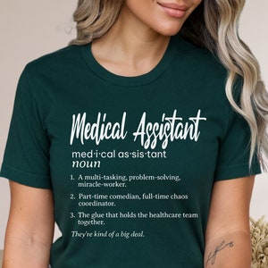 May include: A dark green t-shirt with white text that reads "Medical Assistant" and a definition of the word. The definition includes the phrase "They're kind of a big deal."