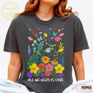 May include: Dark gray t-shirt featuring a colorful floral design with butterflies and the text "ALL WE NEED IS LOVE." The graphic includes flowers in shades of yellow, pink, red, and purple. The shirt is a comfortable, casual style.