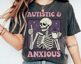Peut inclure: T-shirt gris foncé avec un motif de squelette levant le pouce et tenant une tasse. Le texte "AUTISTIC & ANXIOUS" est en rose, dans un cadre violet. Le t-shirt est de style décontracté.
