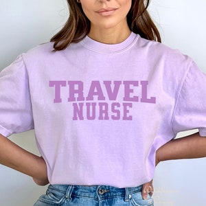 Könnte beinhalten: Ein lavendelfarbenes T-Shirt mit dem Aufdruck "TRAVEL NURSE" in fetter, weißer Schrift.