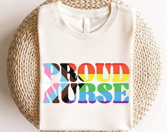 May include: A white t-shirt with the words "PROUD NURSE" in rainbow colors. The letters are outlined in black and filled with different colors of the pride flag.
