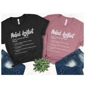 May include: Two t-shirts with the definition of a medical assistant printed on them. The shirts are black and pink and are laid out on a surface with blue jeans and a potted plant.