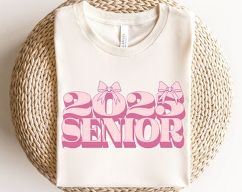 May include: A white t-shirt with a pink graphic that says "2023 Senior" with pink bows on the 2 and 3.