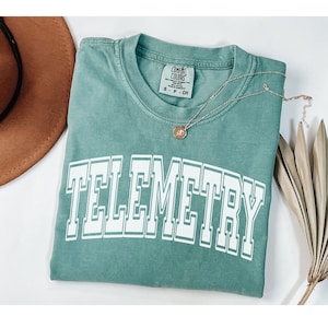 May include: A teal blue t-shirt with the word "TELEMETRY" printed in white with a varsity style font. The shirt is folded and laid flat on a white surface.