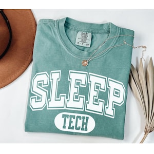 Może przedstawiać: Koszulka w kolorze turkusowym z białym napisem "SLEEP TECH" w stylu uniwersyteckim.
