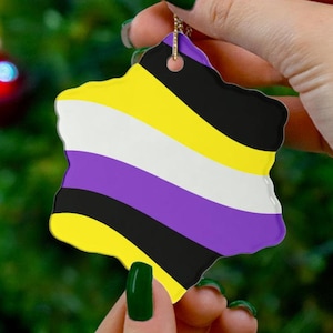 May include: A star-shaped ornament featuring the non-binary pride flag design. The flag has horizontal stripes in yellow, white, purple, and black. The ornament is held by a person with green nail polish, with a Christmas tree in the background.