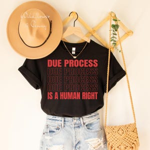 Op de afbeelding: Zwart T-shirt met de tekst "DUE PROCESS" vier keer herhaald in rood, gevolgd door "IS A HUMAN RIGHT" in rood. Het shirt hangt aan een hanger met een strohoed en een stro tas.