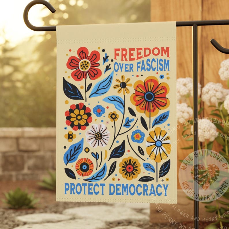 Puede incluir: Una bandera de jard&iacute;n beige con un dise&ntilde;o floral y el texto "FREEDOM OVER FASCISM PROTECT DEMOCRACY" en rojo y azul. La bandera presenta flores y hojas coloridas en tonos de rojo, amarillo, azul y negro.