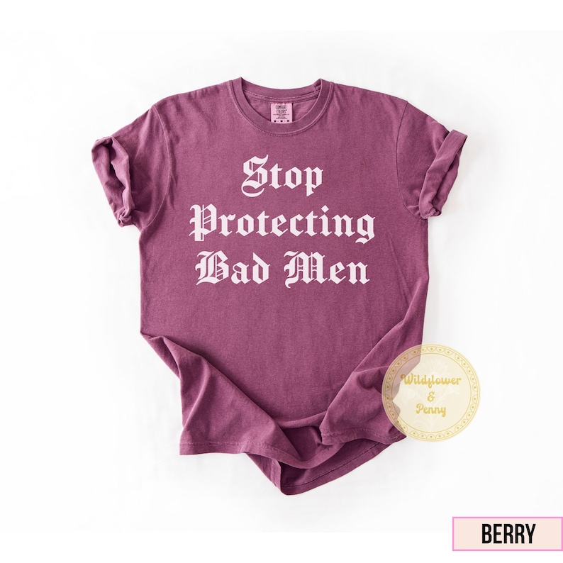 May include: Berry-colored t-shirt with the text "Stop Protecting Bad Men" in a gothic font. The shirt has short sleeves and a crew neck. A small logo is visible on the upper back of the shirt.