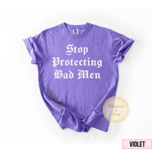 May include: A violet-colored t-shirt with the phrase "Stop Protecting Bad Men" printed in white gothic lettering. The shirt has short sleeves and a crew neck. The shirt is a solid color with a slightly faded appearance.