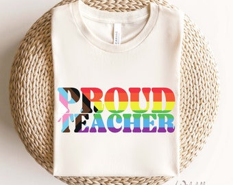 May include: Cream-colored t-shirt with the words "PROUD TEACHER" in rainbow colors and LGBTQ+ flag designs. The shirt is folded on a woven, circular surface. The brand name "Wildflower & Penny" is in the bottom right corner.