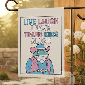 May include: A rectangular garden flag with a white background featuring the text LIVE LAUGH LEAVE TRANS KIDS ALONE in blue and pink. Below the text is an illustration of a frog wearing a pink and blue cowboy hat, vest, and belt.