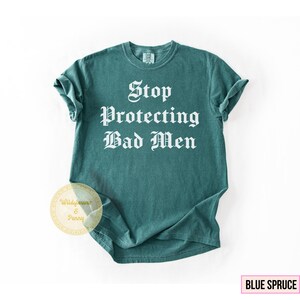 May include: A teal-colored t-shirt with the text "Stop Protecting Bad Men" in a bold, white, gothic font. The shirt has rolled-up sleeves and a crew neck. The words "Blue Spruce" are in a pink box in the lower right corner.