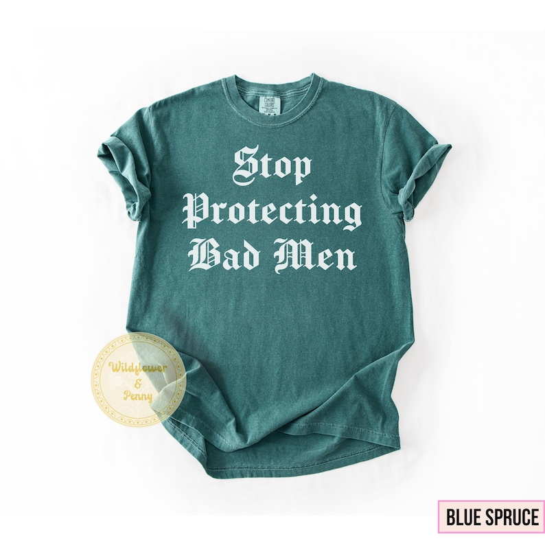 May include: A teal-colored t-shirt with the text "Stop Protecting Bad Men" in a bold, white, gothic font. The shirt has rolled-up sleeves and a crew neck. The words "Blue Spruce" are in a pink box in the lower right corner.