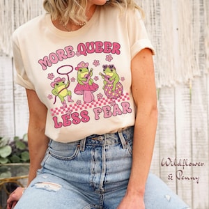 Peut inclure: Un t-shirt cr&egrave;me avec le texte "More Queer Less Fear" en lettres roses. Trois grenouilles de dessin anim&eacute; jouent de la musique sur un fond &agrave; carreaux.
