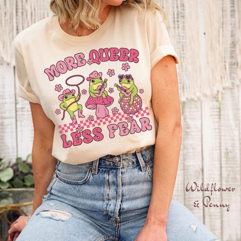 Peut inclure: Un t-shirt cr&egrave;me avec le texte "More Queer Less Fear" en lettres roses. Trois grenouilles de dessin anim&eacute; jouent de la musique sur un fond &agrave; carreaux.