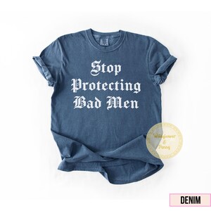 May include: Denim blue t-shirt with the text "Stop Protecting Bad Men" in a gothic font. The shirt has rolled-up sleeves and a round neck. A circular logo with the text "Wildflower & Penny" is on the lower right.