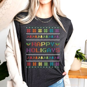 Könnte beinhalten: Ein dunkelgraues T-Shirt mit einem regenbogenfarbenen "Happy Holigays"-Design im Pixel-Stil. Das Design enthält Schneeflocken, Weihnachtsbäume und Rentiere.