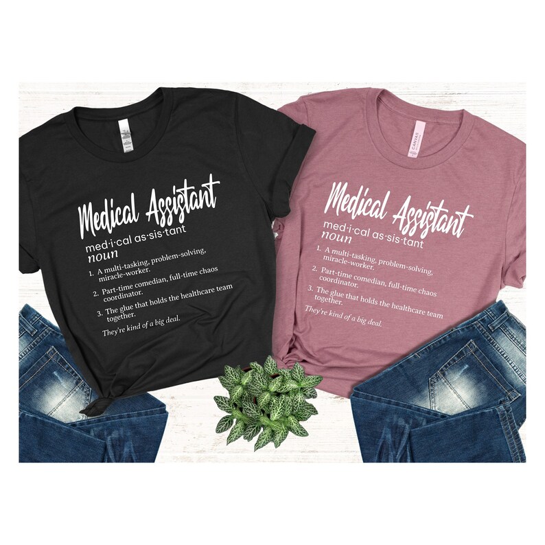 May include: Two t-shirts with the definition of a medical assistant printed on them. The shirts are black and pink and are laid out on a surface with blue jeans and a potted plant.