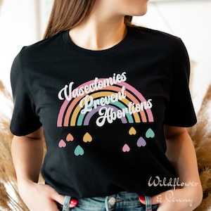 Pu&ograve; includere: Maglietta nera con una grafica arcobaleno e il testo "Vasectomies Prevent Abortions". L'arcobaleno &egrave; composto da giallo, arancione, rosa, viola e verde. Ci sono piccoli cuori di vari colori sotto l'arcobaleno.