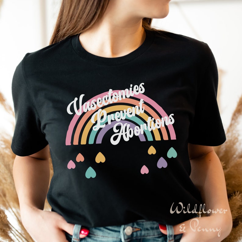 Pu&ograve; includere: Maglietta nera con una grafica arcobaleno e il testo "Vasectomies Prevent Abortions". L'arcobaleno &egrave; composto da giallo, arancione, rosa, viola e verde. Ci sono piccoli cuori di vari colori sotto l'arcobaleno.