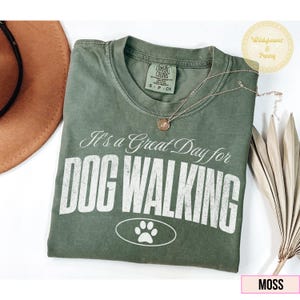 Könnte beinhalten: Ein moosgrünes T-Shirt mit dem weißen Text "It's a Great Day for DOG WALKING". Das Shirt hat eine Pfotenabdruck-Grafik und eine runde Anhänger-Halskette. Ein brauner Hut und eine getrocknete Pflanze sind ebenfalls im Bild.