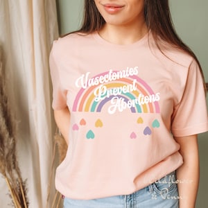Pu&ograve; includere: Una maglietta rosa con un arcobaleno grafico e il testo "Vasectomies Prevent Abortions" in bianco. L'arcobaleno &egrave; composto da diversi colori, tra cui rosso, arancione, giallo, verde, blu e viola. Ci sono piccoli cuori bianchi sparsi intorno all'arcobaleno.