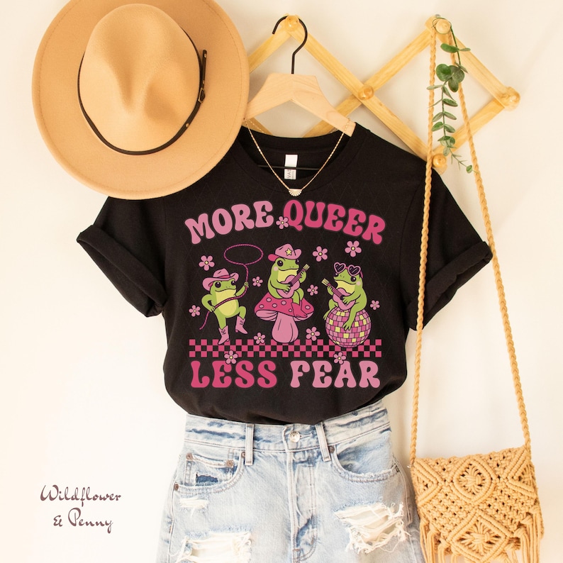 Peut inclure: T-shirt noir avec un motif rose et vert de trois grenouilles jouant de la musique. Le texte "More Queer Less Fear" est imprim&eacute; en lettres roses sur le t-shirt.