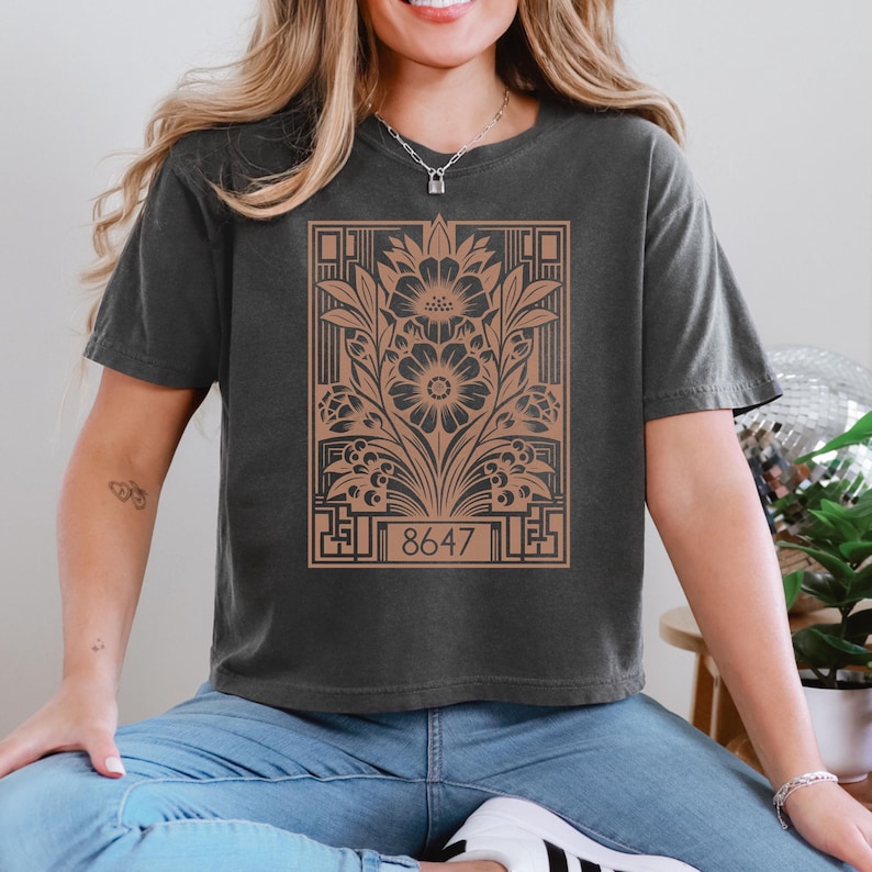 May include: A charcoal gray cropped t-shirt with a brown floral design and the number 8647 printed on the front.