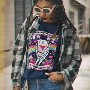 Op de afbeelding: Een marineblauw t-shirt met een afbeelding van een skeletthand die een koffiebeker vasthoudt, omringd door een regenboog, roze bloemen en hartjes. De tekst luidt FUELED BY COFFEE & FEMINIST RAGE. Het shirt wordt gedragen onder een grijs en zwart flanellen overhemd.