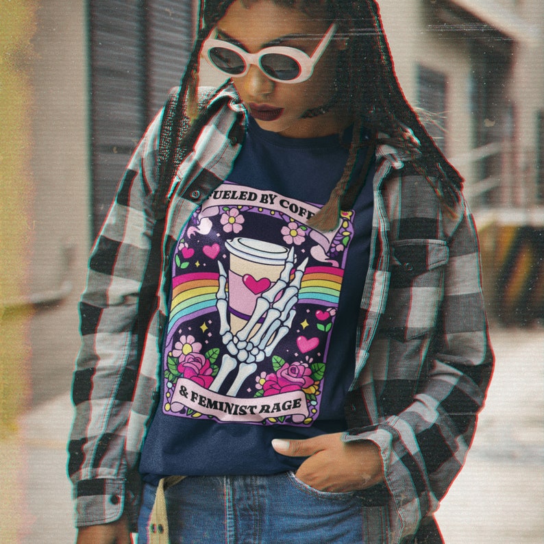 Op de afbeelding: Een marineblauw t-shirt met een afbeelding van een skeletthand die een koffiebeker vasthoudt, omringd door een regenboog, roze bloemen en hartjes. De tekst luidt FUELED BY COFFEE & FEMINIST RAGE. Het shirt wordt gedragen onder een grijs en zwart flanellen overhemd.