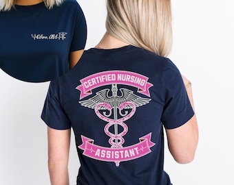 Op de afbeelding: Een marineblauw T-shirt met een roze en zilveren medisch symbool op de rug. Het symbool toont een slang die om een staf met vleugels is gewikkeld. De tekst "CERTIFIED NURSING ASSISTANT" staat op een roze banner onder het symbool.