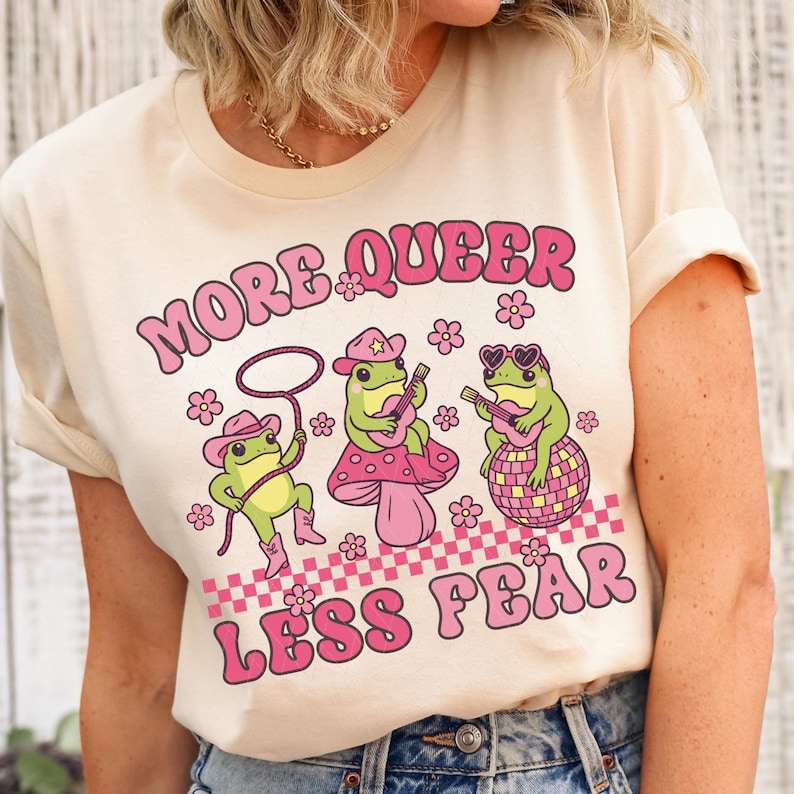 Peut inclure: Un t-shirt cr&egrave;me avec un motif rose et blanc de trois grenouilles jouant de la musique. Le texte "More Queer Less Fear" est imprim&eacute; en lettres roses sur le t-shirt.