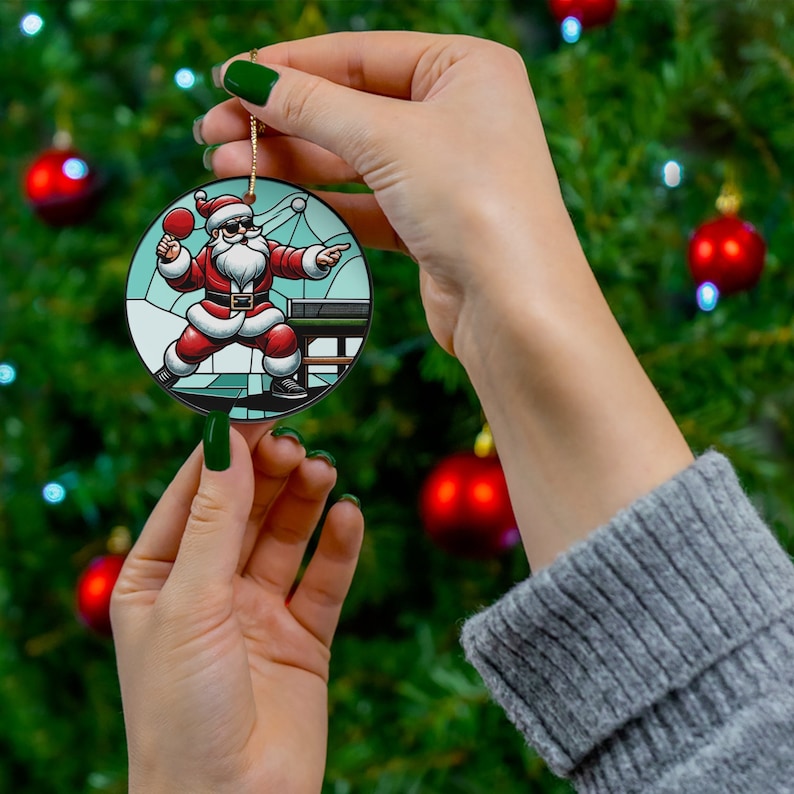 May include: A round Christmas bauble depicting a cartoon illustration of Father Christmas playing table tennis. The bauble has a stained glass effect with a blue, green and white background.