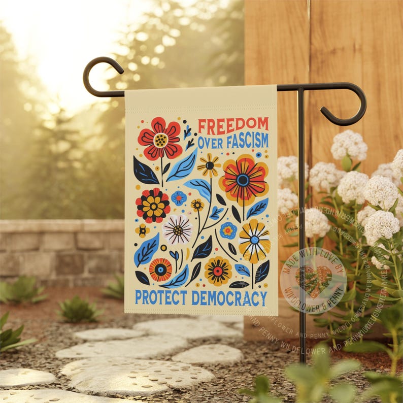 Puede incluir: Una bandera de jard&iacute;n beige con un dise&ntilde;o floral y el texto "FREEDOM OVER FASCISM" y "PROTECT DEMOCRACY" en azul. La bandera presenta flores coloridas en rojo, naranja, amarillo y azul, con hojas verdes.